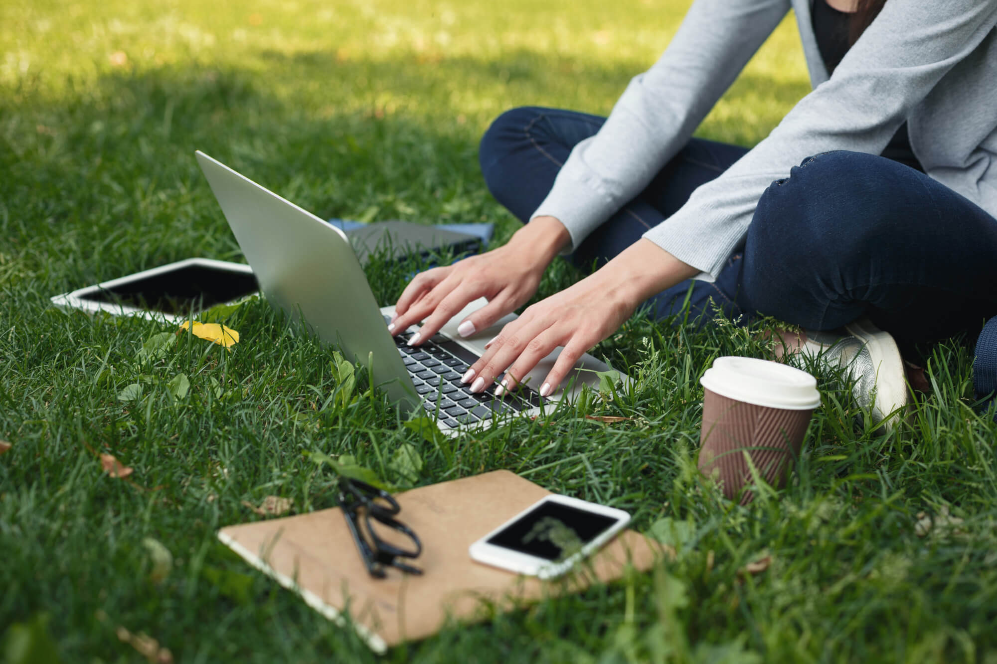 Woman sitting on grass in the shade, using laptop and drinking coffee