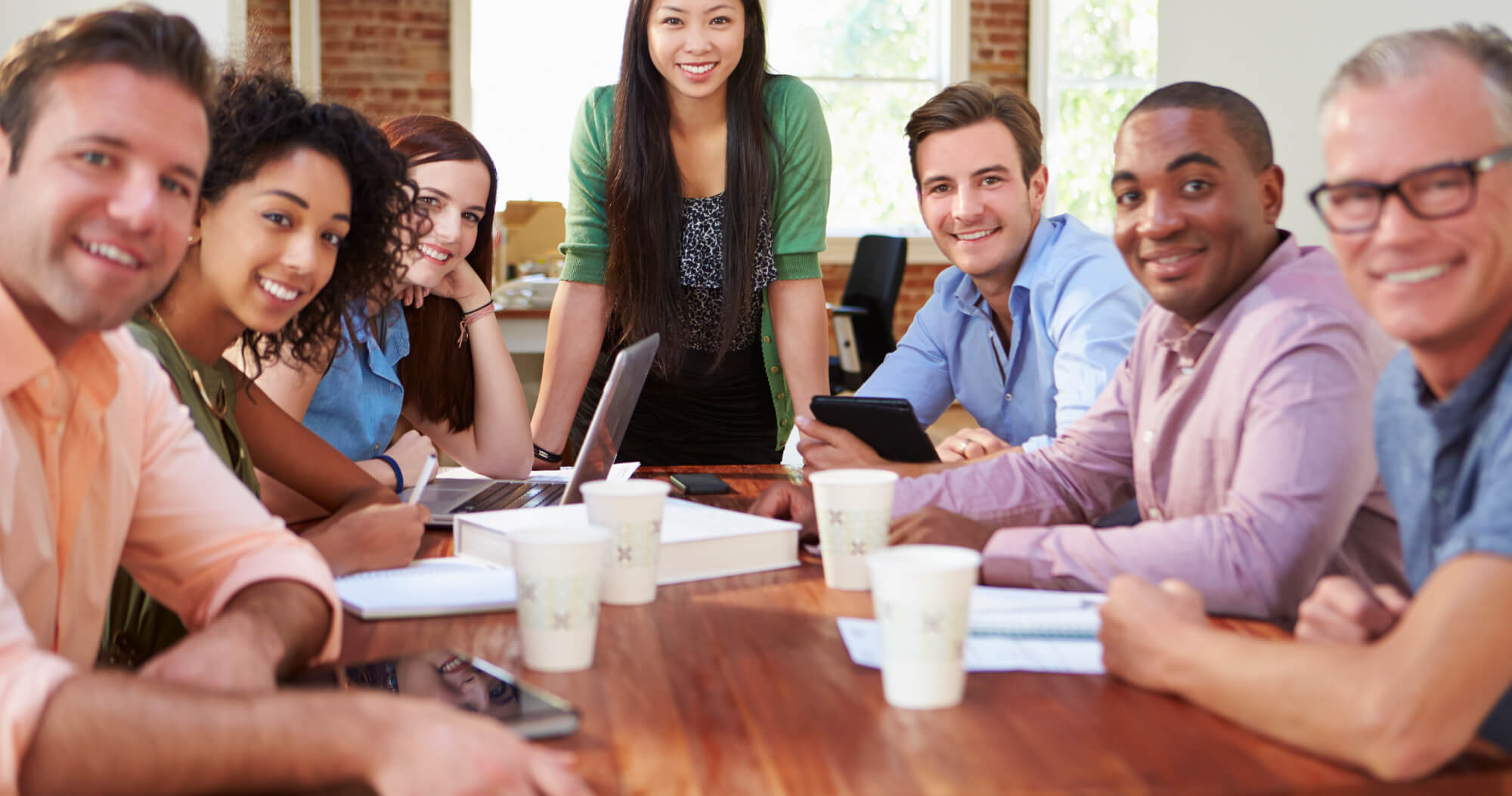 Smiling Asian woman standing at head of table with 6 colleagues looking towards the viewer